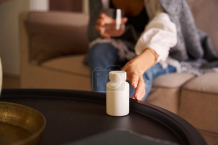 Foto de La mujer se sienta con un vaso de agua en el sofá, se acerca a la mesa para tomar una botella de pastillas - Imagen libre de derechos