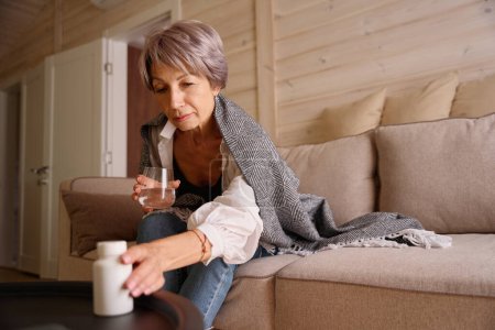 Téléchargez les photos : Malade pensionné s'assoit avec un verre d'eau et pot de pilules sur le canapé, elle est enveloppée dans un châle chaud - en image libre de droit