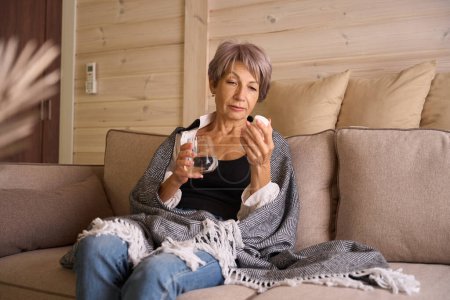 Photo for Sick woman sits with a glass of water and a jar of pills on the sofa, she is wrapped in a warm shawl - Royalty Free Image