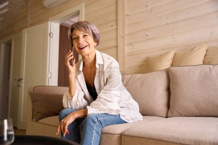 Foto de Señora sonriente en ropa casual se comunica en su teléfono móvil, en una habitación con un interior minimalista - Imagen libre de derechos