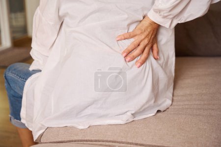 Téléchargez les photos : Femme malade souffre de maux de dos, elle est assise sur le canapé à la maison - en image libre de droit