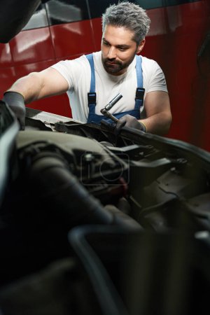 Téléchargez les photos : Sérieux concentré caucasien technicien de service automobile vérifier sur les pièces de voiture sous le capot - en image libre de droit