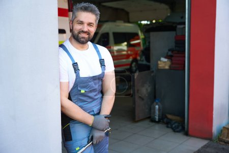 Foto de Mecánico de coche sonriente con llave en la mano apoyado contra la pared en el taller de reparación de automóviles - Imagen libre de derechos