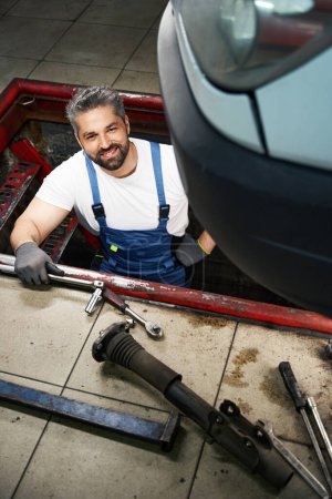 Photo for Pleased car mechanic with wrench in his hand standing on stairs of vehicle inspection pit - Royalty Free Image