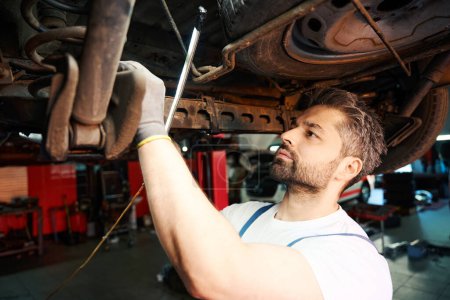 Téléchargez les photos : Travailleur d'atelier de réparation automobile concentré sérieux inspectant les pièces de suspension de véhicule avec la lampe - en image libre de droit