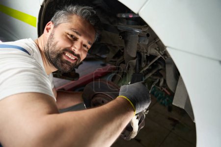 Foto de Sonriente técnico de servicio automotriz que mide la velocidad de rotación del disco de la rueda del automóvil con tacómetro - Imagen libre de derechos