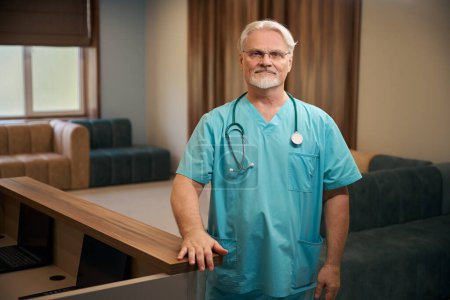 Calm physician with stethoscope around neck leaning on reception desk in medical facility lobby