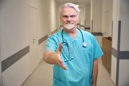 Téléchargez les photos : Portrait taille haute d'un travailleur de la santé avec stéthoscope autour du cou, main tendue pour serrer la main alors qu'il se tenait debout dans le couloir de l'hôpital - en image libre de droit