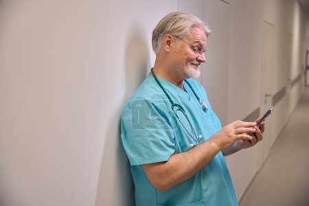Téléchargez les photos : Travailleur médical concentré appuyé contre le mur dans le couloir de l'hôpital tout en lisant quelque chose sur l'écran du smartphone - en image libre de droit