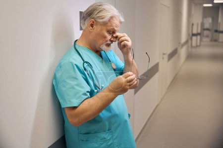 Téléchargez les photos : Clinicien épuisé avec des lunettes à la main pincer son nez pont tout en s'appuyant contre le mur dans le couloir de l'hôpital - en image libre de droit