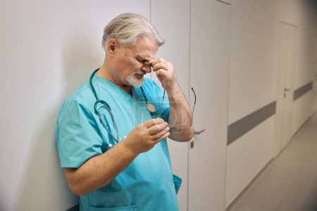 Téléchargez les photos : Médecin fatigué avec des lunettes à la main pincer son nez pont tout en s'appuyant contre le mur dans le couloir de l'hôpital - en image libre de droit