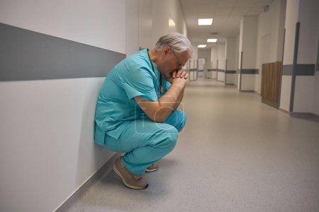 Téléchargez les photos : Vue latérale de clinicien coûteux assis sur ses hanches près du mur dans le couloir de l'hôpital - en image libre de droit