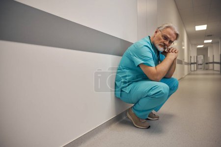 Téléchargez les photos : Travailleur de la santé assis sur ses hanches près du mur dans le couloir de l'hôpital - en image libre de droit