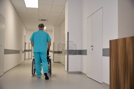 Téléchargez les photos : Vue arrière du travailleur de la santé poussant une chaise roulante par des poignées le long d'un couloir d'hôpital vide - en image libre de droit