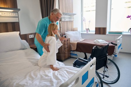 Téléchargez les photos : Travailleur de la santé soulevant une patiente handicapée du lit d'hôpital au fauteuil roulant dans la salle - en image libre de droit