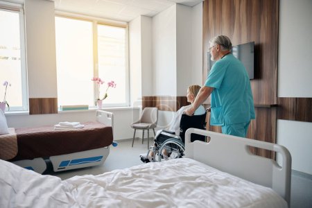Téléchargez les photos : Fauteuil roulant roulant de travailleur médical avec femme hospitalisée vers fenêtre dans la chambre d'hôpital - en image libre de droit