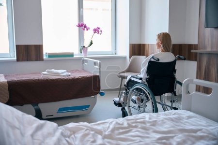 Photo for Back view of woman with disability sitting in wheelchair in front of hospital room window - Royalty Free Image