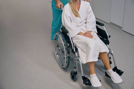 Téléchargez les photos : Photo recadrée d'une infirmière poussant une chaise roulante avec un patient le long du couloir dans un établissement pour malades hospitalisés - en image libre de droit