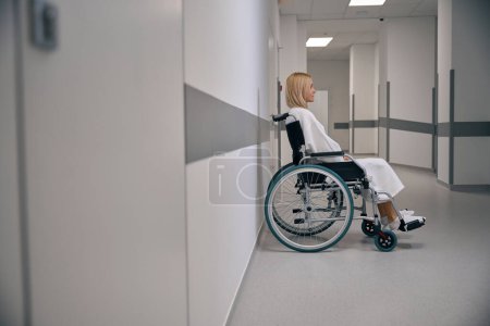 Téléchargez les photos : Vue latérale d'une femme hospitalisée ayant une déficience assise dans un fauteuil roulant dans un couloir d'un établissement médical - en image libre de droit