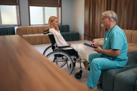 Photo for Clinician taking notes on clipboard while looking at hospitalized woman seated in wheelchair in medical facility lobby - Royalty Free Image