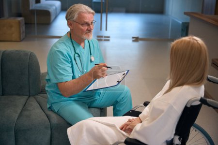 Téléchargez les photos : Médecin masculin avec stylo et presse-papiers assis sur le canapé communiquant avec une patiente hospitalisée en fauteuil roulant dans le foyer de l'établissement de soins de santé - en image libre de droit
