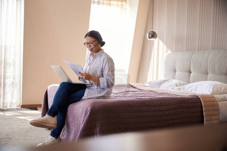 Foto de Mujer de negocios con auriculares y portátil sentado en la cama en suite mirando a través de documentos en papel - Imagen libre de derechos