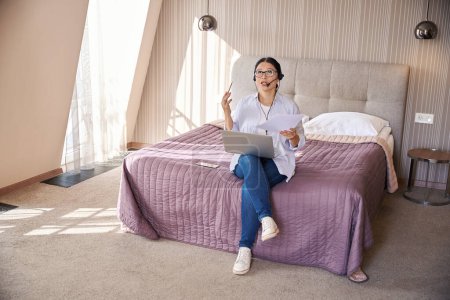 Photo for Business lady seated on bed with computer and documents talking into headset microphone during Internet phone call in hotel room - Royalty Free Image