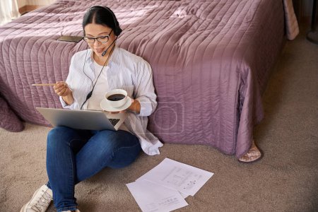 Photo for Teleworker with headset seated on carpeted floor in suite with cup of espresso and pencil watching something on computer monitor - Royalty Free Image