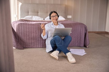 Foto de Freelancer sonriente con portátil sentado en el suelo alfombrado en la habitación de hotel hablando en el micrófono de auriculares durante la llamada telefónica a Internet - Imagen libre de derechos
