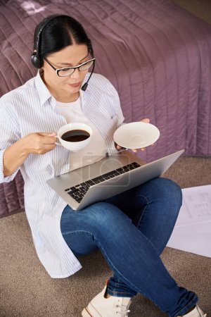 Téléchargez les photos : Travailleur à distance sérieux avec ordinateur portable assis sur le tapis dans la chambre pendant la pause café - en image libre de droit