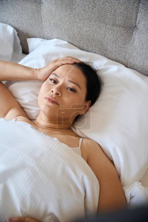 Téléchargez les photos : Triste femme touchant sa tête avec la main alors qu'elle était couchée dans son lit enveloppée dans une couverture - en image libre de droit