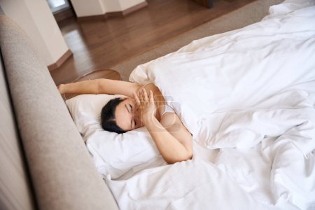 Foto de Mujer bostezando envuelta en una manta acostada sobre una almohada en una cómoda cama en su suite - Imagen libre de derechos