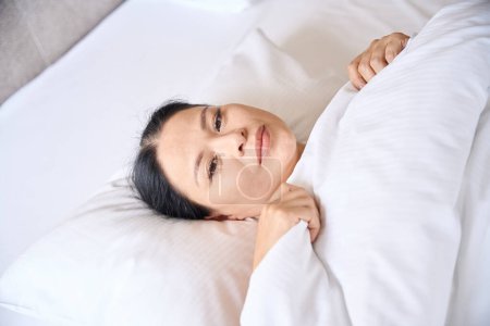 Téléchargez les photos : Femme mature sereine tirant la couverture sur elle-même tout en étant couché sur un oreiller doux - en image libre de droit