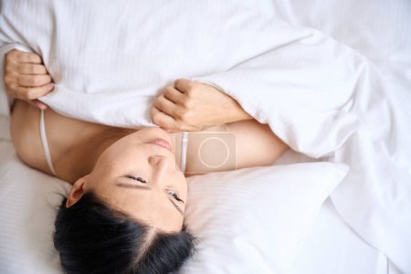Foto de Calma hembra tirando de edredón sobre sí misma mientras está acostada sobre suave almohada blanca - Imagen libre de derechos