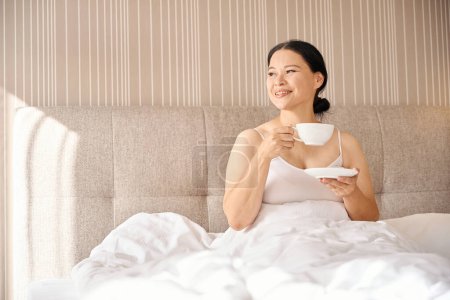 Téléchargez les photos : Femme souriante avec tasse de café et soucoupe dans les mains assises dans le lit en suite et regardant loin - en image libre de droit