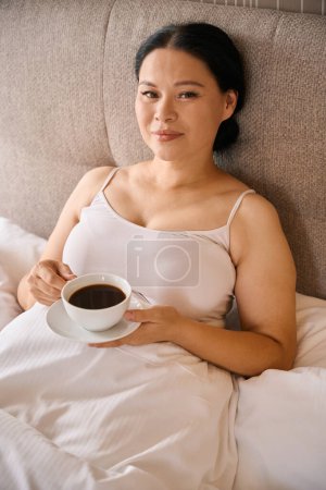 Téléchargez les photos : Dame agréable avec tasse de café et soucoupe dans les mains assises dans le lit après le réveil - en image libre de droit
