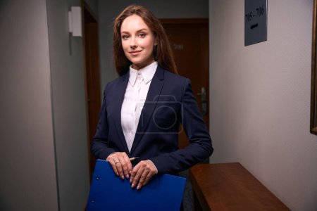 Photo for Contented housekeeping supervisor with clipboard and pen in hands standing in hotel corridor - Royalty Free Image