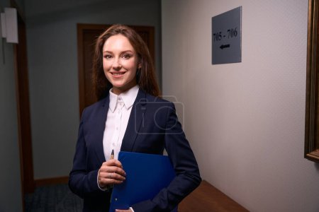 Photo for Waist-up portrait of cheerful manager with pen and clipboard in her hands standing in hotel hallway - Royalty Free Image