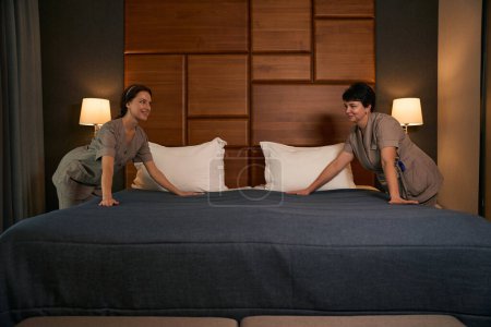 Photo for Smiling maid and her female colleague smoothing out surface of bedspread with hands during bed-making in guest suite - Royalty Free Image