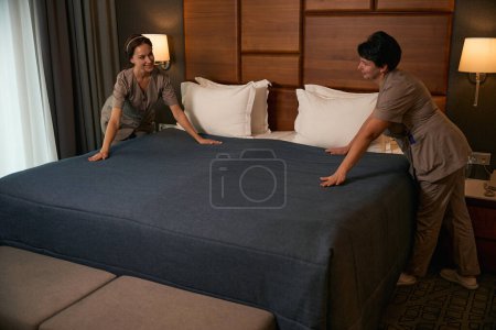 Photo for Joyful chambermaid smoothing out bedspread surface with hands during bed-making assisted by female colleague - Royalty Free Image