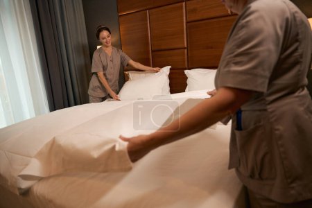 Photo for Smiling chambermaid in uniform and her colleague making bed in guest suite - Royalty Free Image