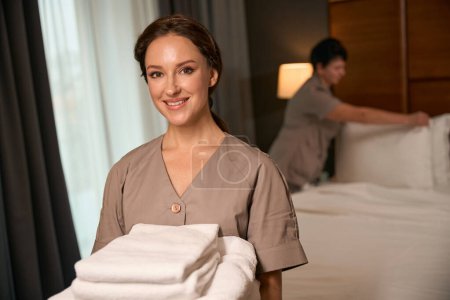 Photo for Joyous maid with stack of clean towels in hands posing for camera while her colleague making bed in guest suite - Royalty Free Image