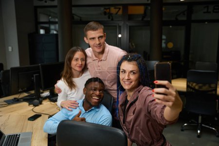 Foto de Hombres y mujeres multiétnicos reunidos cerca del lugar de trabajo en una cómoda oficina y haciendo selfies, celebrando el aniversario de la compañía, posando ante la cámara y sonriendo, alegre equipo amigable - Imagen libre de derechos