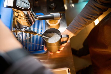 Foto de Cafetera irreconocible o barman batiendo leche para latte en jarra de acero en la cafetería - Imagen libre de derechos