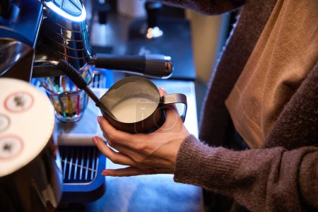 Foto de Mujer desconocida camarera barista batiendo leche para latte en jarra de acero en la cafetería - Imagen libre de derechos