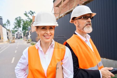 Photo for Pleased construction manager with document folder and foreman with walkie-talkie standing together on road among half-built houses - Royalty Free Image