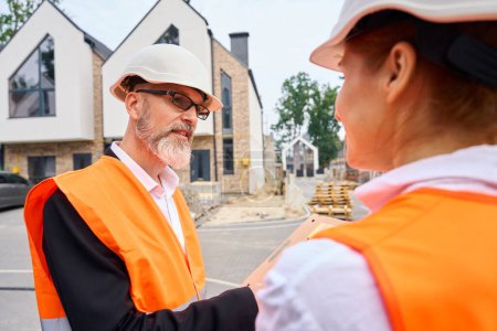 Photo for Serious foreman with open document folder in hands talking to his female colleague in front of half-built house - Royalty Free Image
