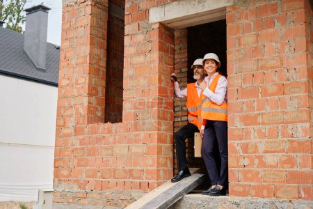 Photo for Smiling female building inspector and foreman standing in doorway of half-built house and looking at something into distance - Royalty Free Image