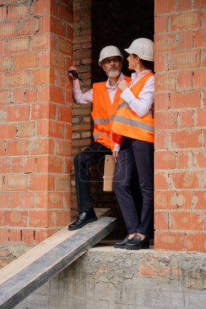 Foto de Capataz serio con walkie-talkie en la mano hablando con el inspector del edificio en la puerta de la casa inacabada - Imagen libre de derechos
