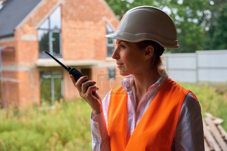 Photo for Portrait of female construction supervisor talking on walkie-talkie while standing in front of unfinished house - Royalty Free Image